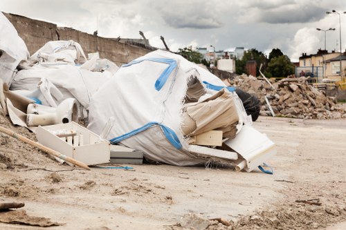 A large pile of discarded construction and household waste materials is situated on an unpaved outdoor surface, with various white and beige plastic items, fabric, and debris visible. The pile includes rolled-up coverings, plastic piping, and foam or insulation materials, all loosely arranged and partially spilling onto the ground. In the background, there is a partially demolished or under-construction area with dirt, rubble, and a retaining wall made of soil or materials, along with distant buildings and trees under a cloudy sky. This scene is typical of rubbish left for removal or clearance on a site requiring professional waste management, as provided by Commercial Waste Removal Crouch End, serving areas near N8 postcode, with a focus on efficient rubbish collection and disposal services.