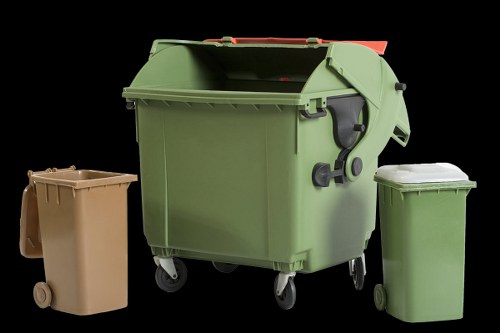 The image displays three wheelie bins positioned on a dark background. The largest bin, centrally located, is green with a textured surface, black handles, and a closed lid, indicating it is used for general rubbish collection. To its left, there is a smaller beige bin with a smooth surface and solid wheels, also with a closed lid, typically used for household waste. On the right side of the green bin, there is another small green bin with a matching textured surface and a closed lid, often used for recycling or specific waste streams. The arrangement suggests a refuse collection setup, with the green bin likely associated with commercial waste removal services in an area such as Crouch End or nearby London. The lighting is even, emphasizing the textures and colours of the bins, which appear to be made from durable plastic with a matte finish. This composition is clearly relevant to rubbish removal and waste management, supporting local SEO for commercial waste services in the Crouch End area.