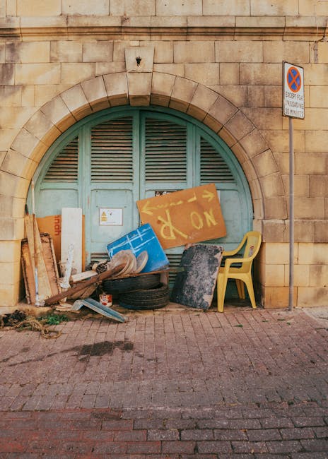 A collection of discarded items is piled against a light blue, arched wooden door set within a stone brick wall. The rubbish includes various materials such as a yellow plastic chair, a black rubber t