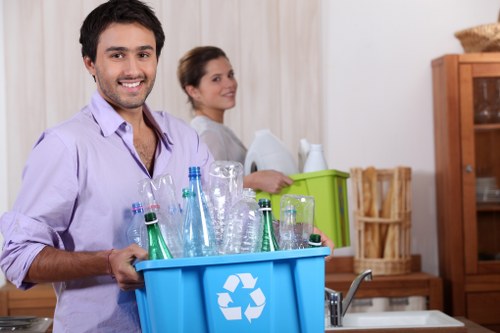 A young man with dark hair, beard, and casual shirt is smiling and holding a blue recycling bin filled with glass bottles and jars, positioned in a kitchen setting. Behind him, a woman with brown hair tied back, wearing a light-colored top, is standing near a counter, with a green plastic container and a wooden chopping board in the background. The kitchen features wooden cabinets and a neutral-toned wall, creating a domestic environment suitable for waste collection and recycling activities. The image emphasizes proper rubbish segregation in a home context, with a focus on recyclable glass items, supporting waste management services such as those offered by Commercial Waste Removal Crouch End. The scene is well-lit with natural or ambient indoor lighting, highlighting the textures and colors of the bottles, containers, and kitchen furnishings, which are typical for a UK household involved in responsible waste disposal practices near N8 postcode area.