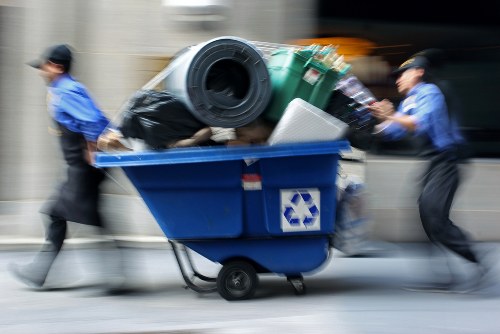 A blue recycling bin mounted on a wheeled trolley, filled with various types of waste including black trash bags, a green plastic container, and a white item, is being pushed by two individuals wearing blue uniforms and black hats along a city street. The background shows a blurred urban scene with a vehicle in motion, indicating activity around a commercial or residential area in Crouch End. The scene illustrates rubbish collection or disposal activities typical for local waste management services, with a focus on waste removal for businesses or residents. The bin's surface appears slightly textured, and the waste materials are stacked in a somewhat disorganized manner, with the green container and black bags visibly protruding. The lighting suggests daytime with natural light illuminating the scene, emphasizing practical waste management operations in the local area.