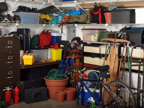 The image depicts a cluttered storage area or garage filled with various household and outdoor items. In the foreground, there are multiple plastic flower pots of different sizes, some stacked inside each other, and a coiled garden hose resting in a large terracotta pot. To the right, a blue bicycle with a black saddle and red accents leans against a shelving unit. The shelving unit is filled with assorted objects, including boxes, toolcases, and containers, with a variety of miscellaneous items such as a black toolbox, a red and black bag, and a yellow plastic container. Above, there are metal or wire shelving units holding more plastic storage boxes, bags, and outdoor equipment, including a yellow extension cord, a pair of black work boots, and a red and black backpack. The environment appears to be a garage or utility space with typical clutter associated with rubbish or waste accumulation, naturally supporting the services provided by Commercial Waste Removal Crouch End in managing refuse and clutter efficiently, especially in the local area around N8 or N19 postcode areas near Crouch End.